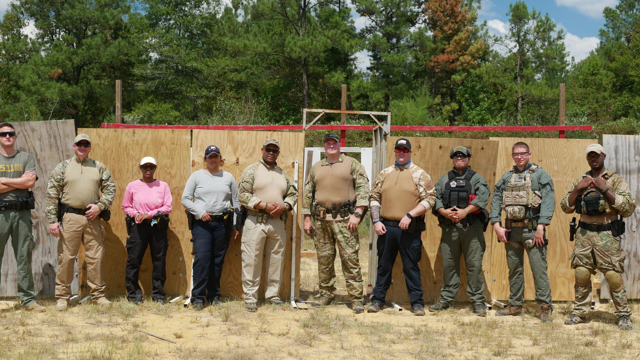 A group of nine people in tactical gear stand in front of wooden structures in a forested outdoor setting, conveying a sense of readiness and teamwork.