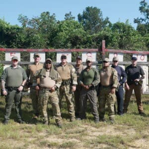 A group of twelve men in tactical and casual attire stand in a line on a grassy field.