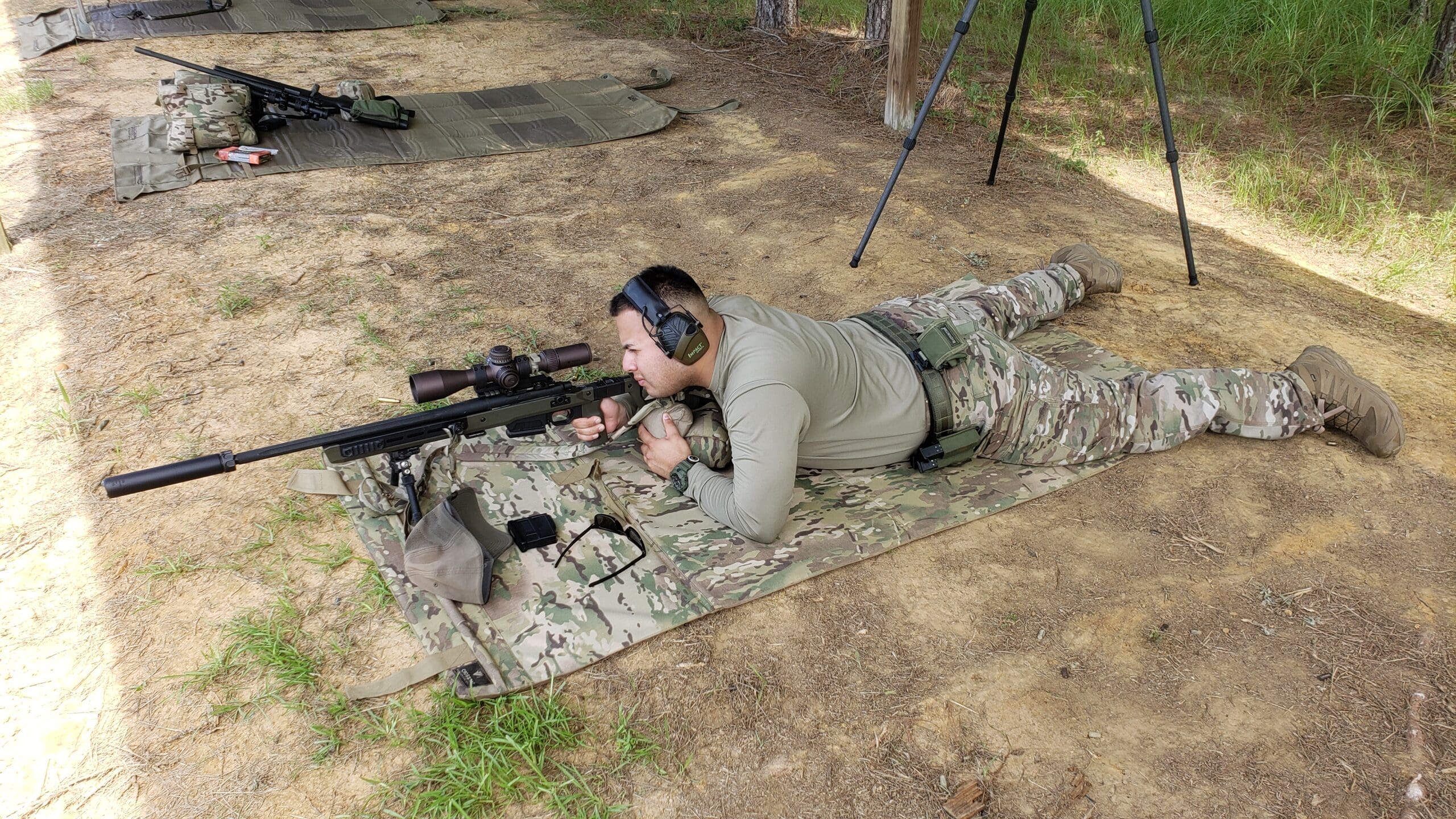 A person in camouflage, lying prone on a mat, aims a rifle at an outdoor shooting range.