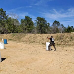 A person with a handgun practices shooting behind cover at a training range, surrounded by barrels and greenery under a clear blue sky.