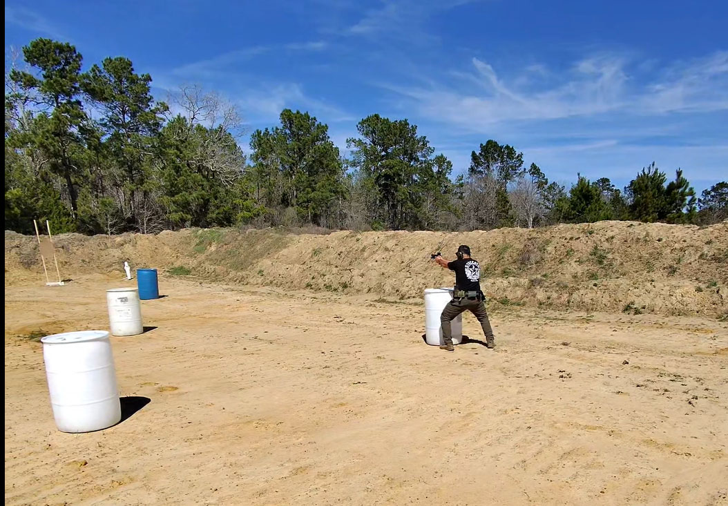 A person with a handgun practices shooting behind cover at a training range, surrounded by barrels and greenery under a clear blue sky.