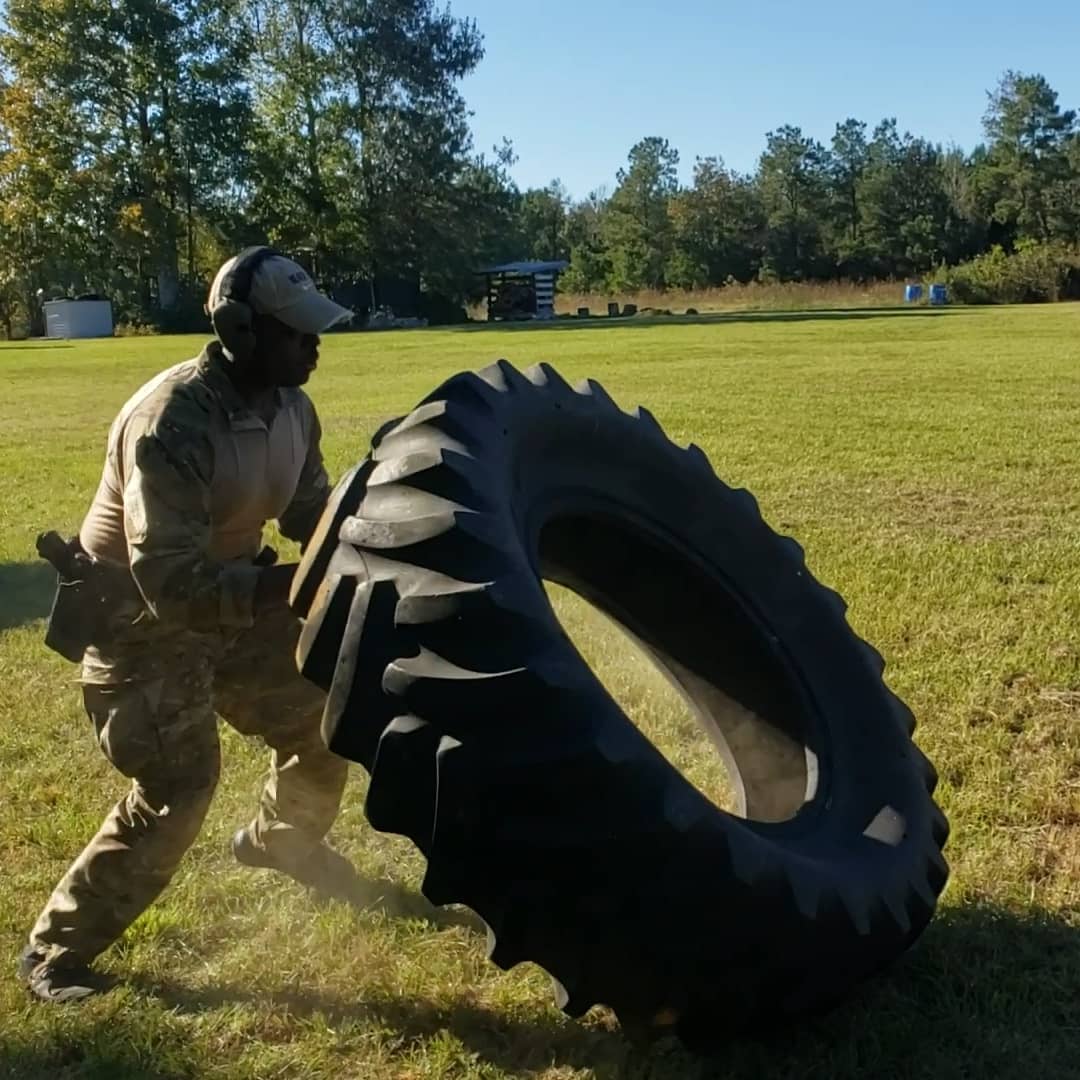 A person in camouflage gear is flipping a large tire on a grassy field, surrounded by trees under a clear blue sky, conveying strength and focus.