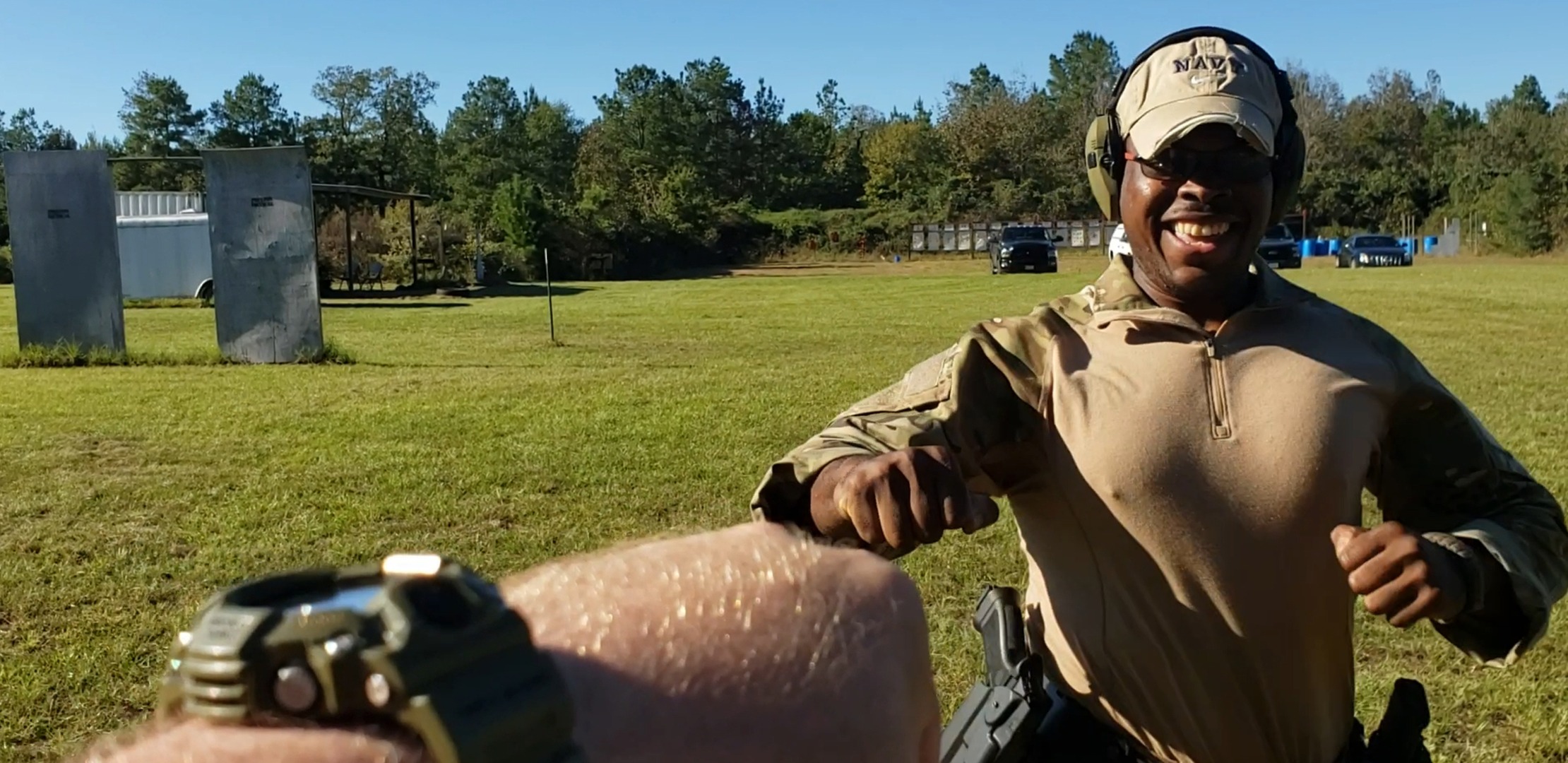 A person in tactical gear and cap, smiling energetically on a sunny shooting range.