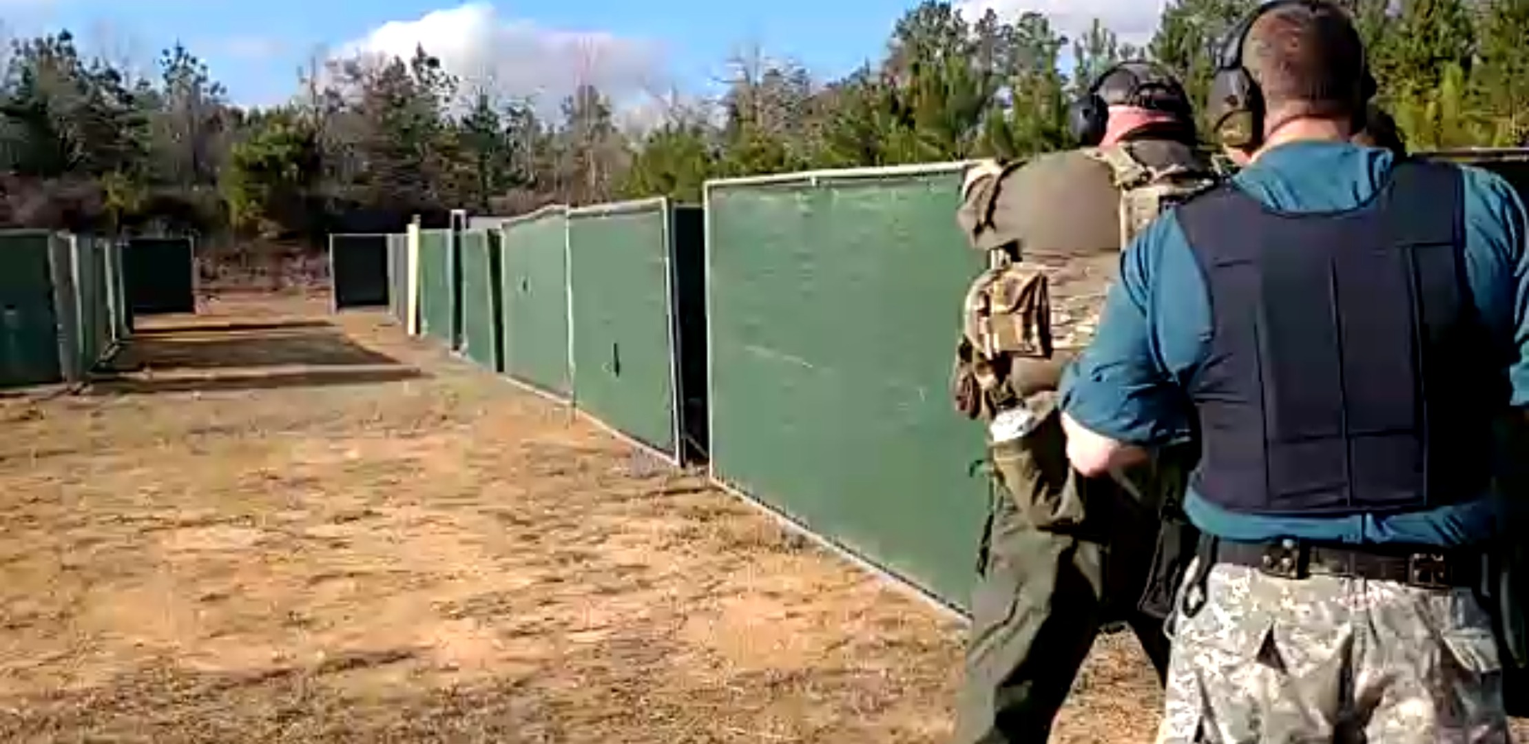 Two people in tactical gear and headphones are at an outdoor shooting range with high green barriers.